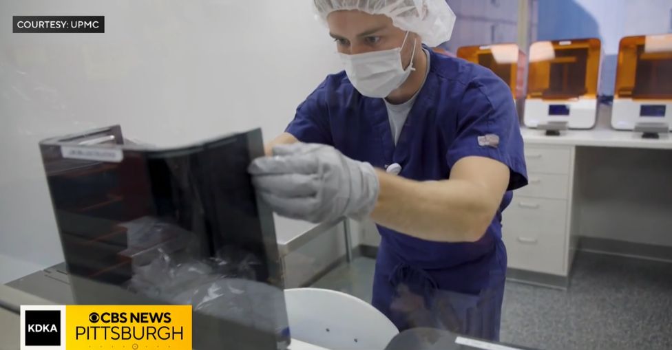 An engineer in scrubs and a hair covering lifting a printed part from the tank of a 3D printer in the UPMC lab